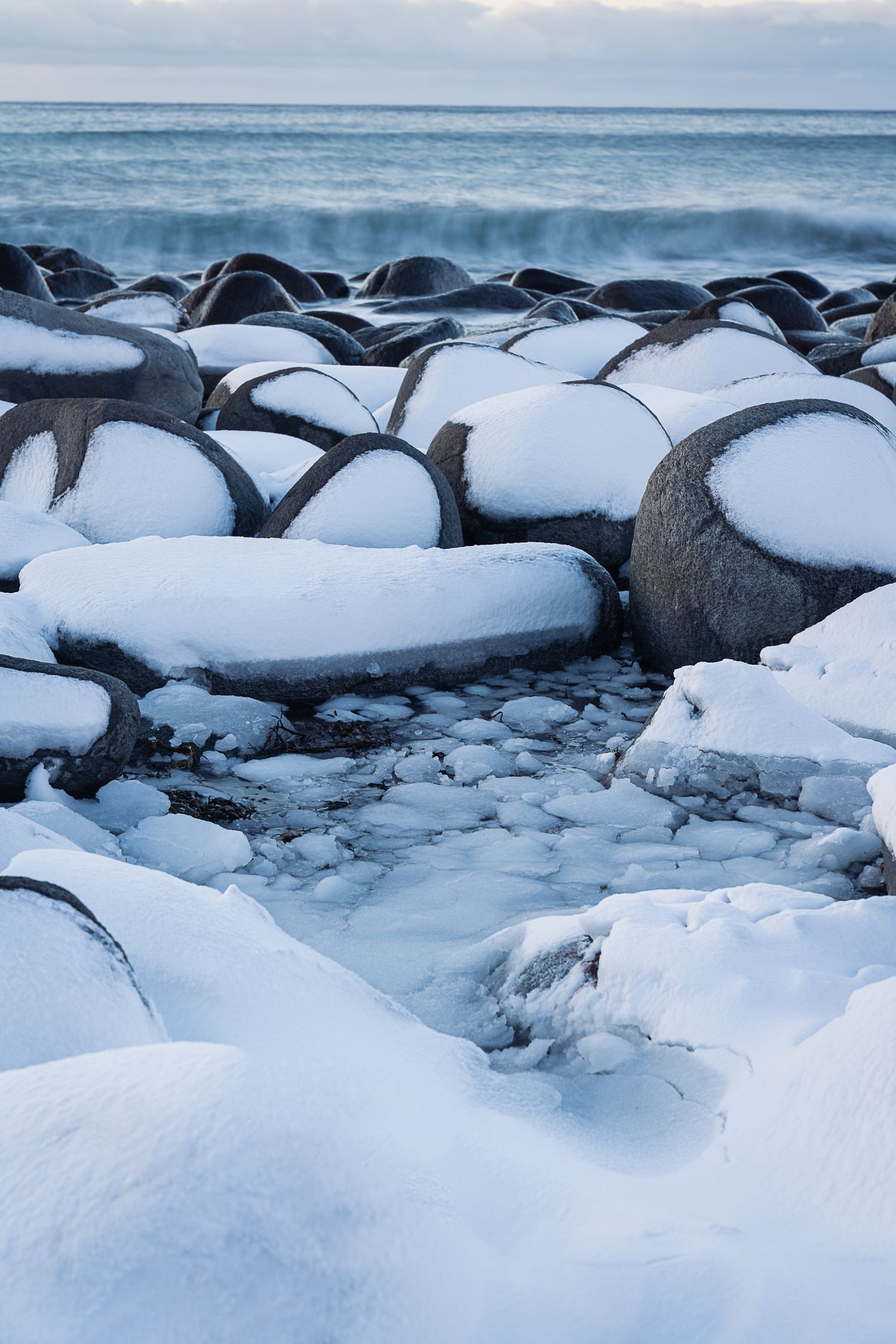 Snow-covered boulders line a rocky winter shoreline with waves crashing in the background under a cloudy sky.