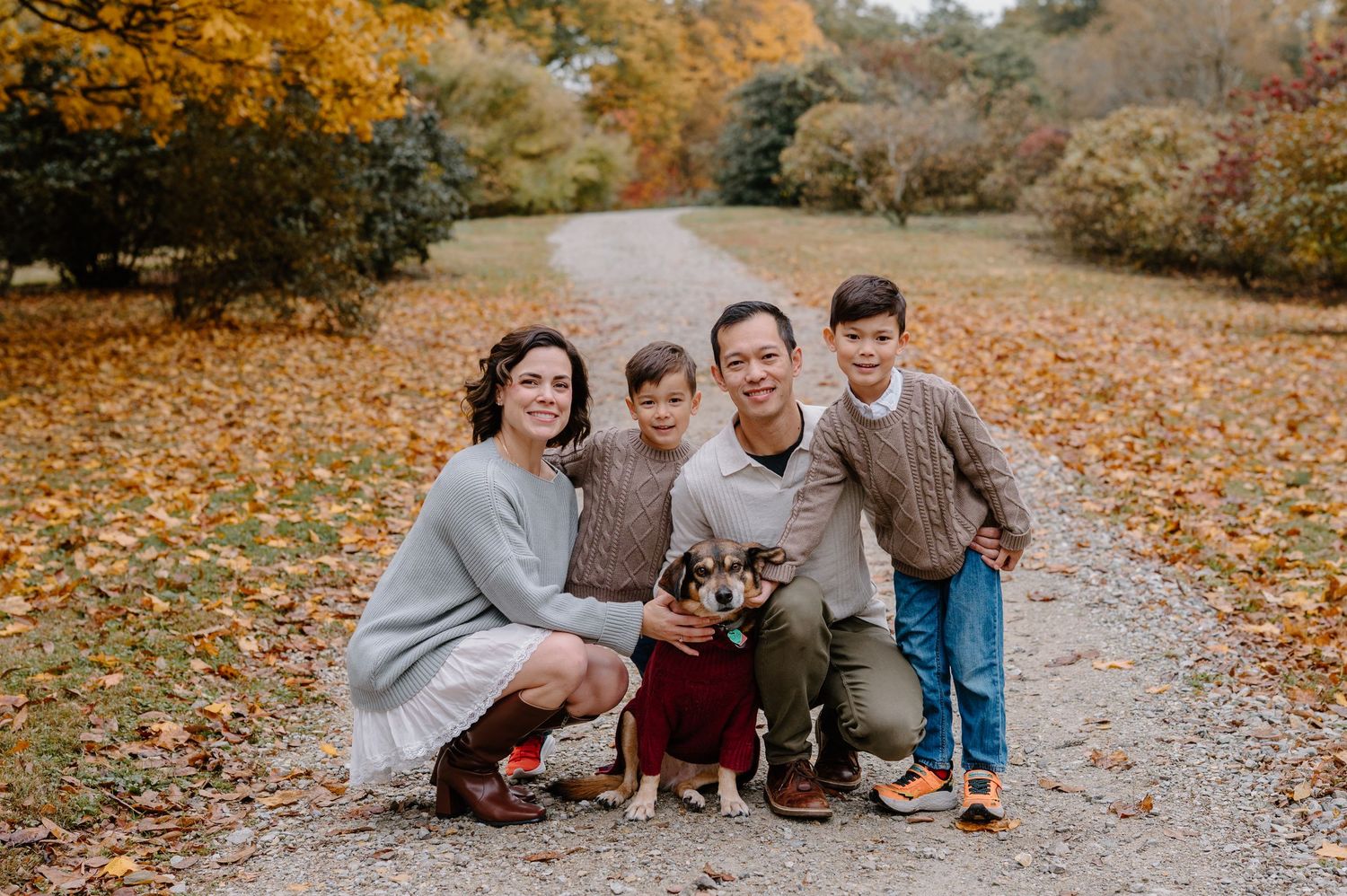 Family of four smiling outdoors in autumn park with dog, surrounded by colorful fall foliage on a gravel path.