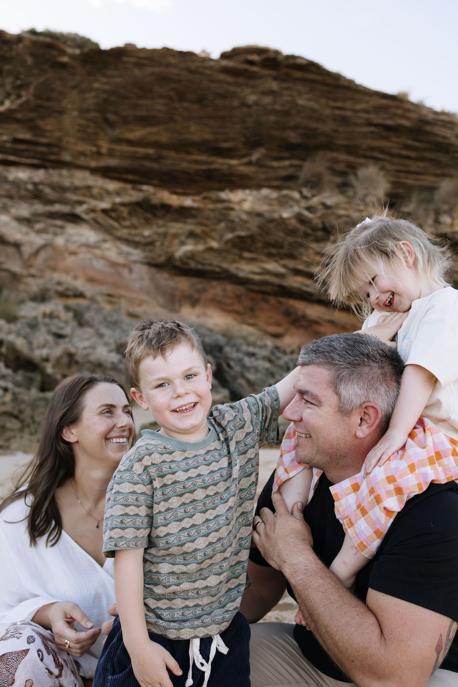 Happy family of four laughing together outdoors near rocky cliffs, parents with two young children enjoying a sunny day.