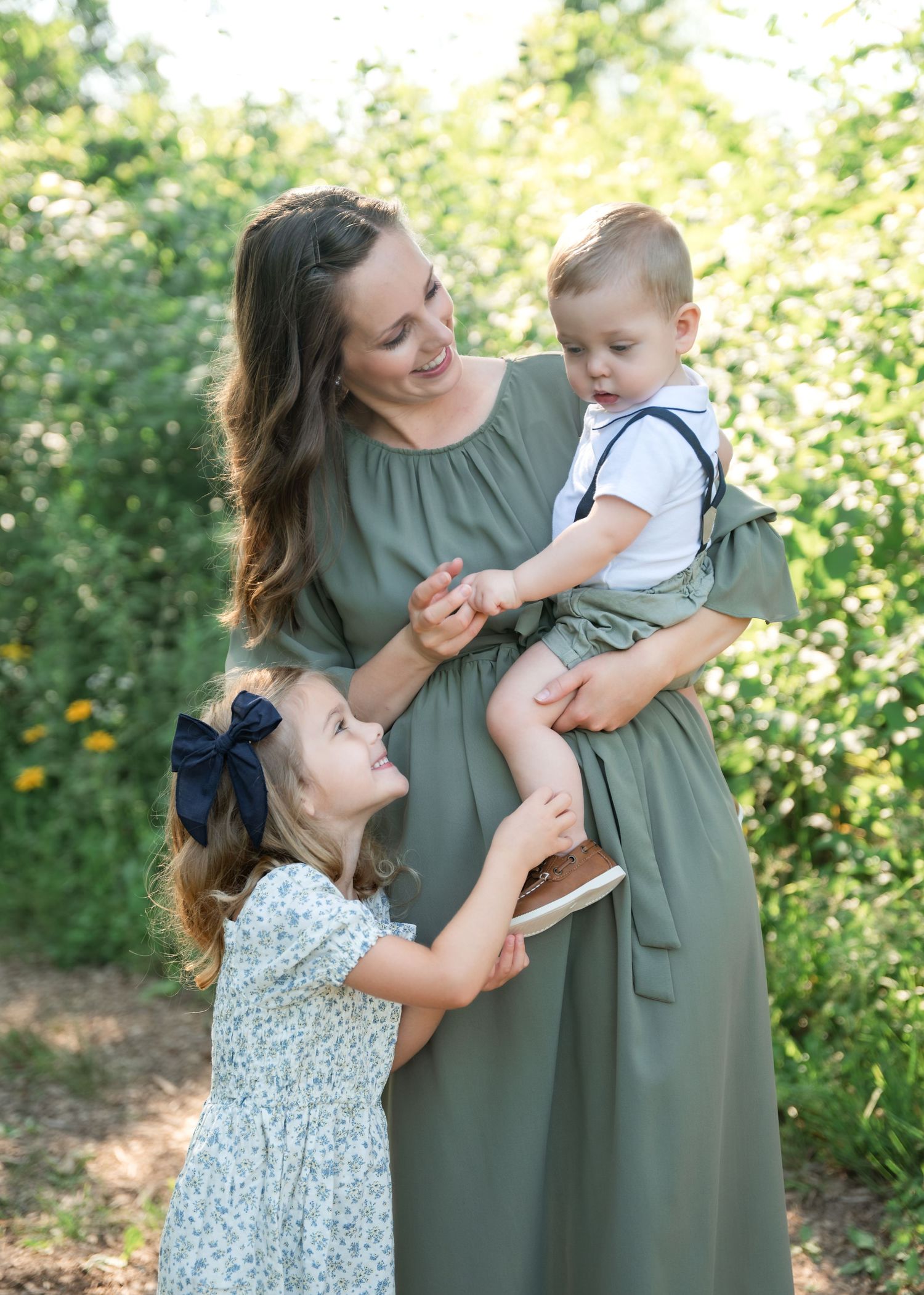 Pregnant woman in olive dress holds toddler boy while young girl in floral dress looks up smiling outdoors.