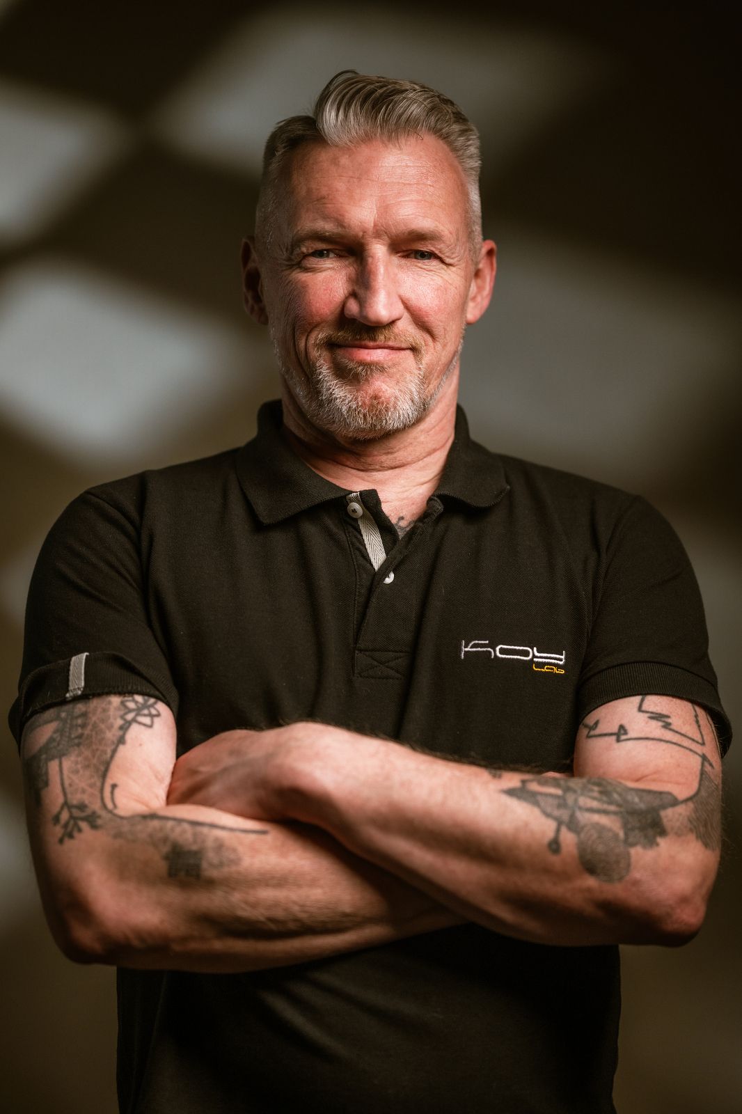 Professioneel zelfportret van fotograaf Bart Boodts met grijs haar en een korte baard, in een zwarte polo en met gekruiste, getatoeëerde arm
