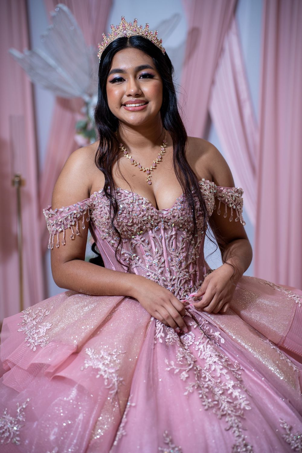 Smiling young woman wearing pink quinceañera dress with lace bodice, gold tiara, and necklace indoors.