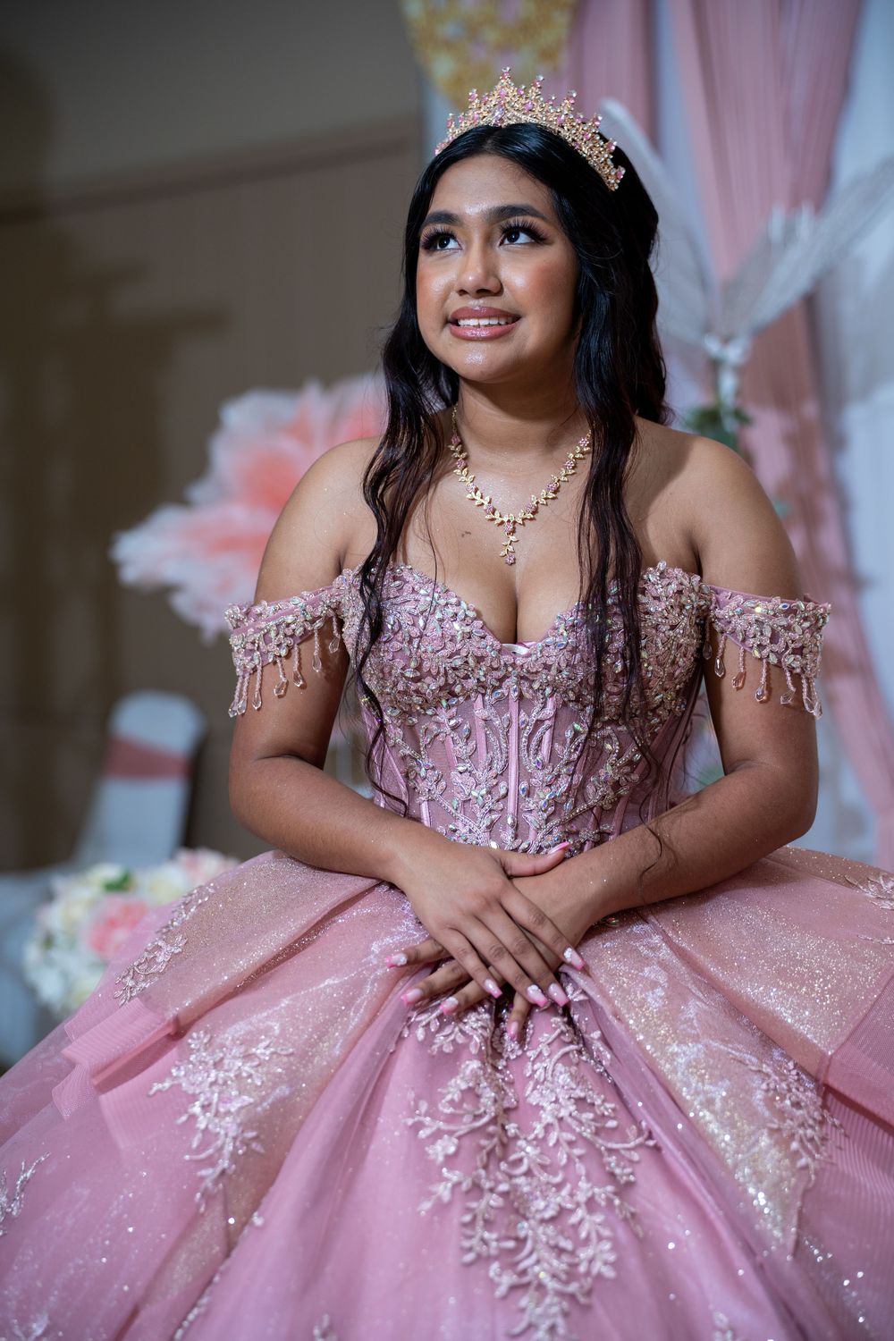Latina quinceañera in pink lace ball gown and crown smiles while posing at her celebration venue.