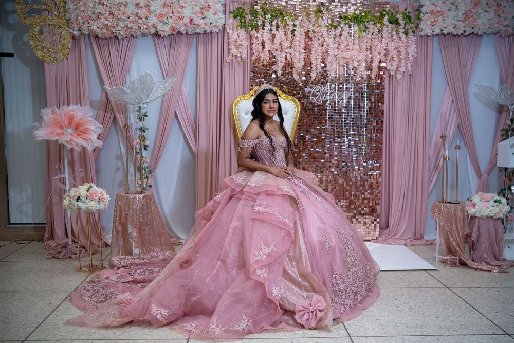 Quinceañera girl in pink ball gown sits on gold throne surrounded by pink floral decorations.