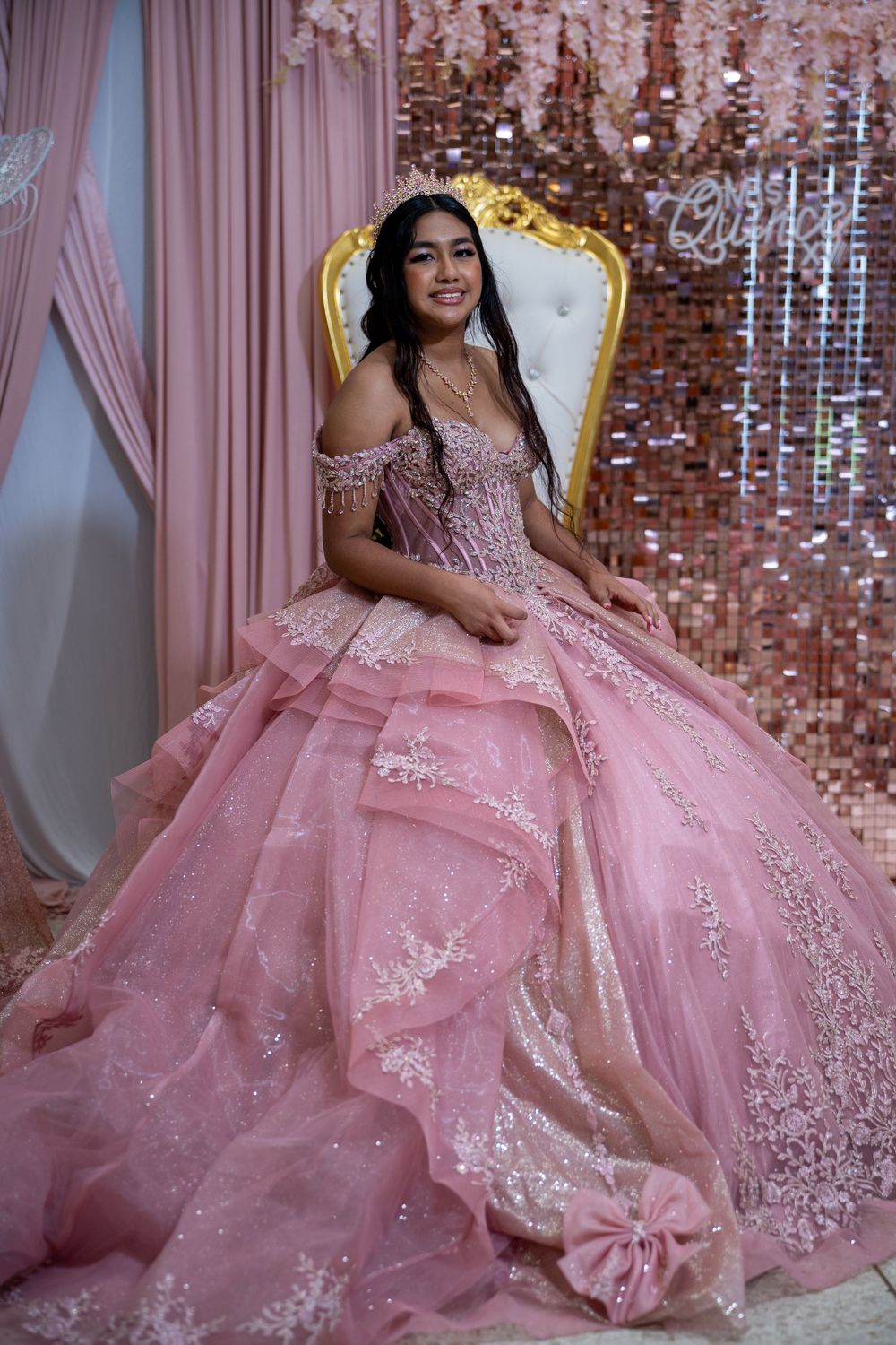 Smiling quinceañera in pink lace ball gown poses on ornate gold and white throne.