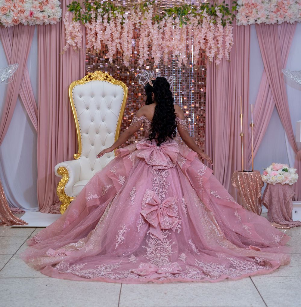 Back view of quinceañera in pink ball gown with large bow train sitting on white tufted throne chair.