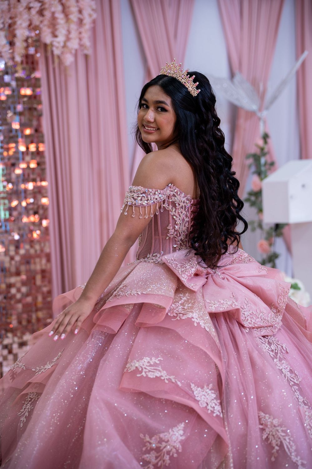 Teen in pink off-shoulder quinceañera gown with bow and crown poses indoors.