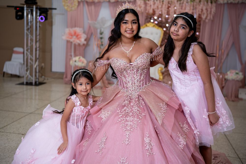 Three girls in pink dresses pose at a quinceañera, with the birthday girl wearing a crown and ornate ball gown.