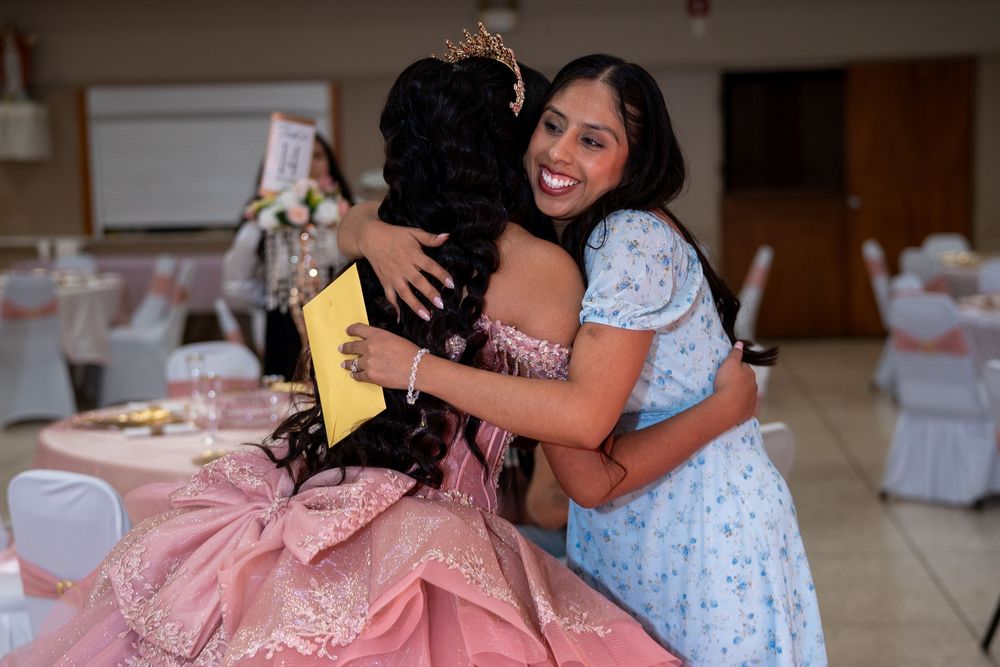 Girl in blue dress hugs quinceañera celebrant in pink ballgown at reception.