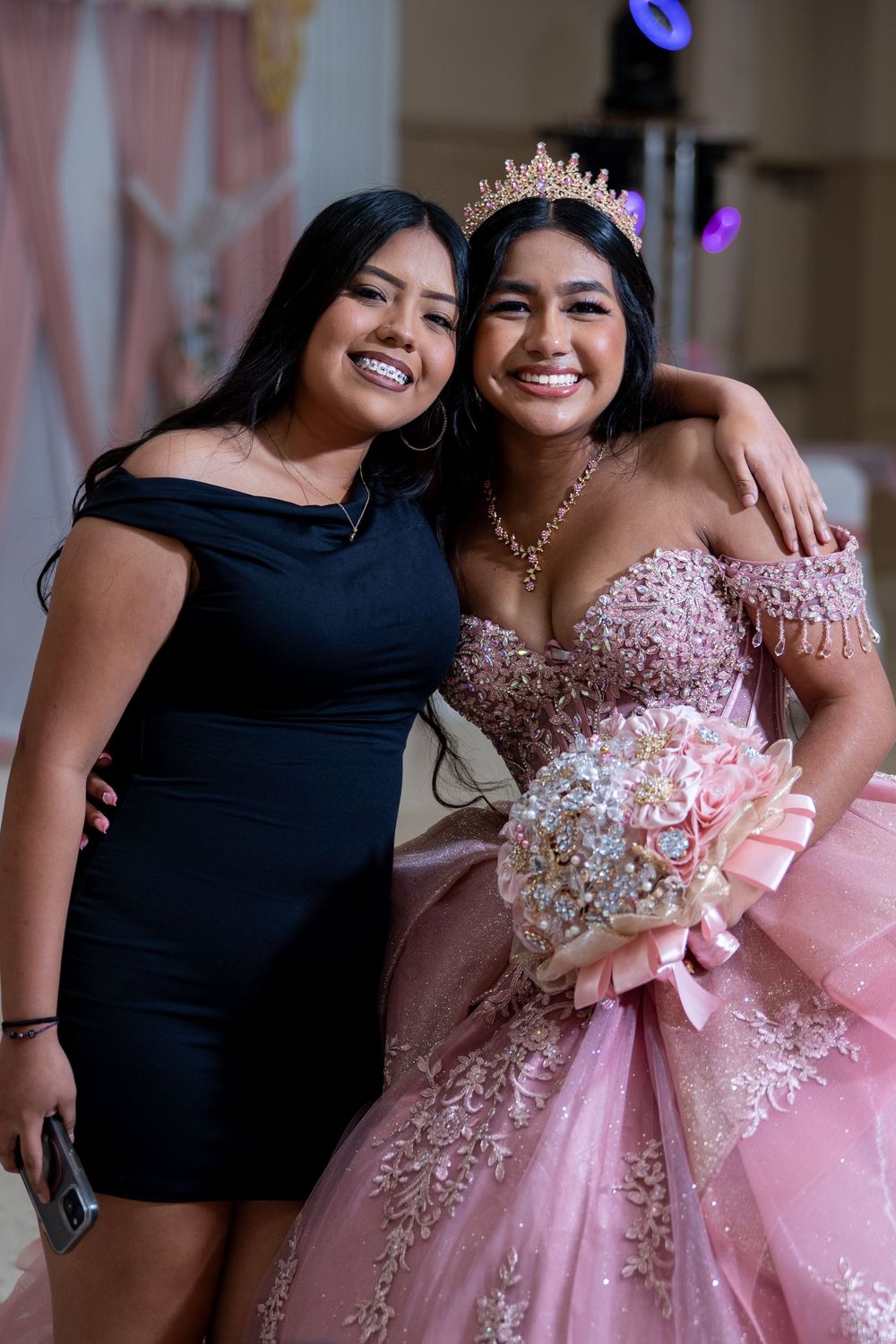 Two young women smile together at a quinceañera, one in a black dress and one in a pink gown with a crown.