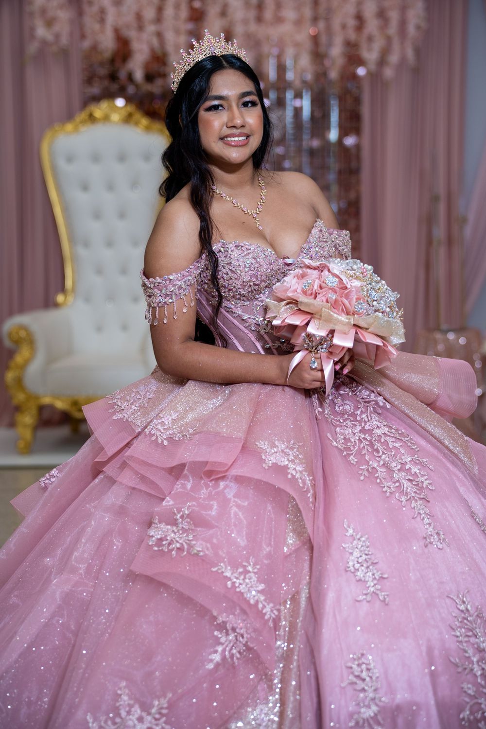 Young woman in pink quinceañera ball gown holds floral bouquet while seated on ornate white throne.