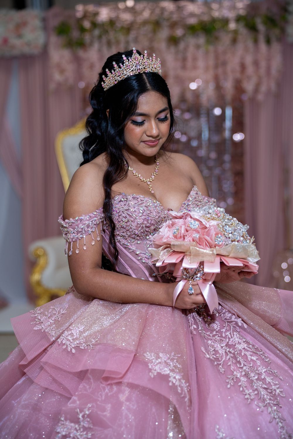Young woman in pink quinceañera gown with tiara holding floral bouquet while looking down gracefully.