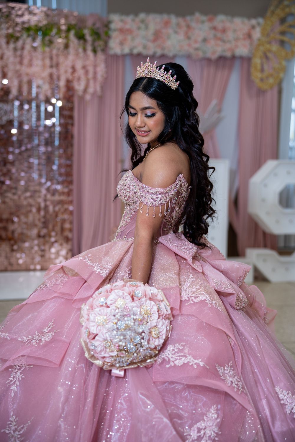 Young woman in pink ball gown with lace, crown tiara, holding floral bouquet at quinceañera celebration.