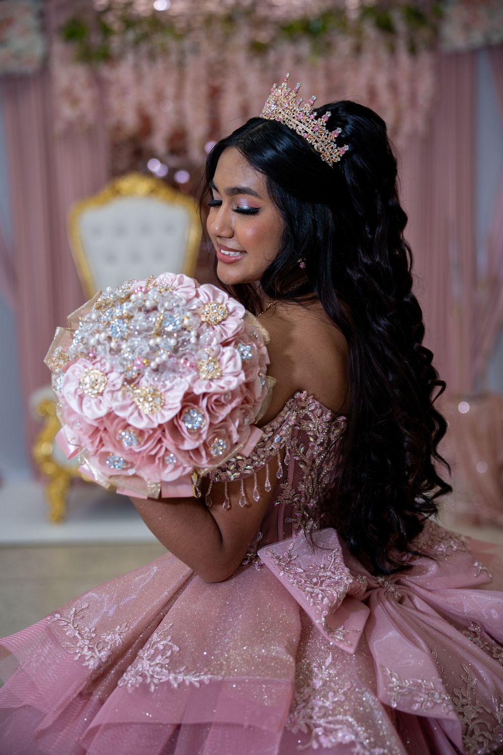 Latina quinceañera in pink gown and crown holds jeweled floral bouquet indoors.