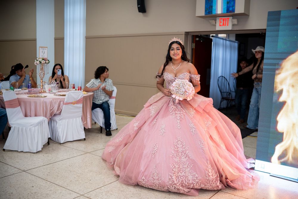 Quinceañera girl in pink ballgown walks into reception hall holding bouquet.