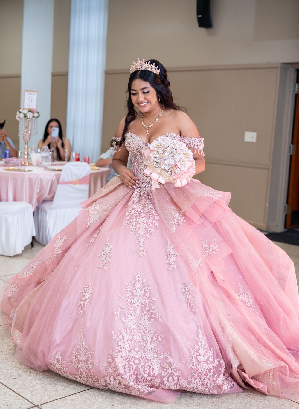 Teen in pink off-shoulder quinceañera ball gown with lace appliqués carrying bouquet at celebration venue.