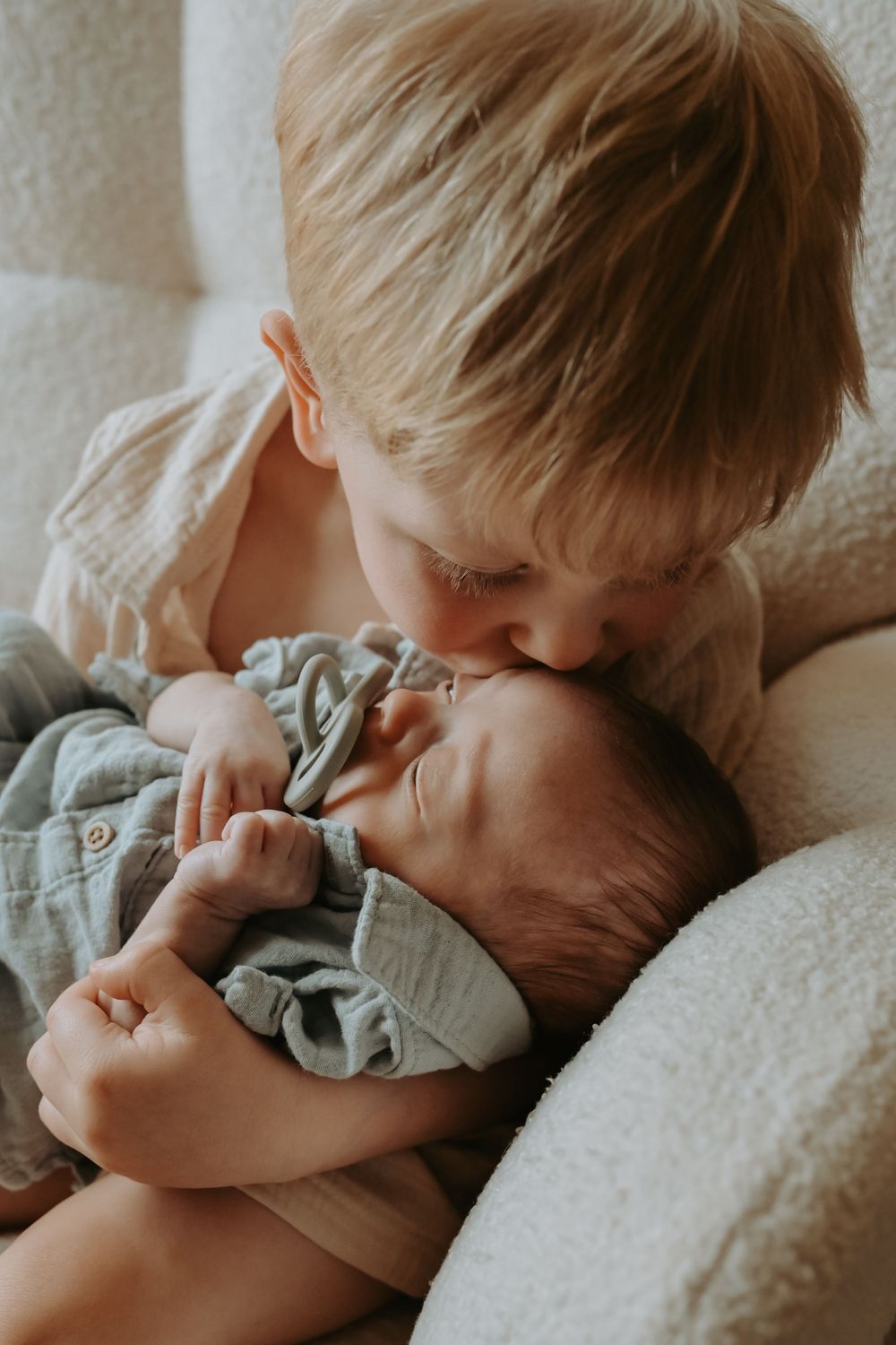 Blonde toddler boy gently kissing newborn baby sibling while holding them in a cozy white chair.