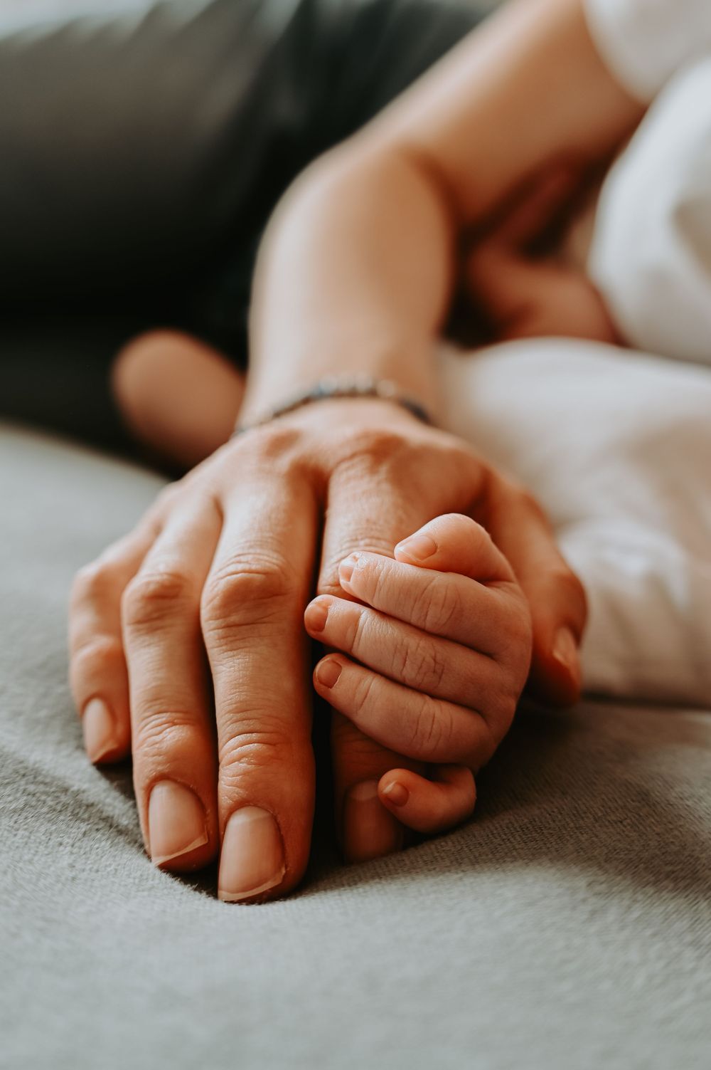 A tender close-up of an adult hand gently holding a tiny newborn baby's hand on a soft gray surface.