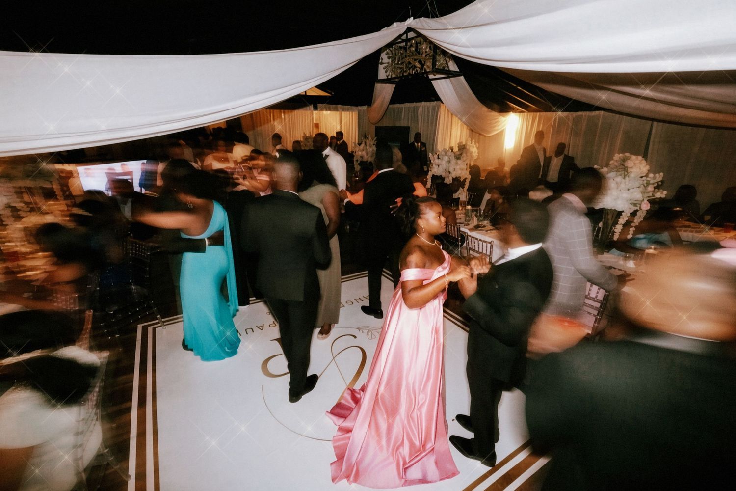 Bride in a pink gown dances on a white dance floor surrounded by wedding guests under draped ceiling fabric at the reception.