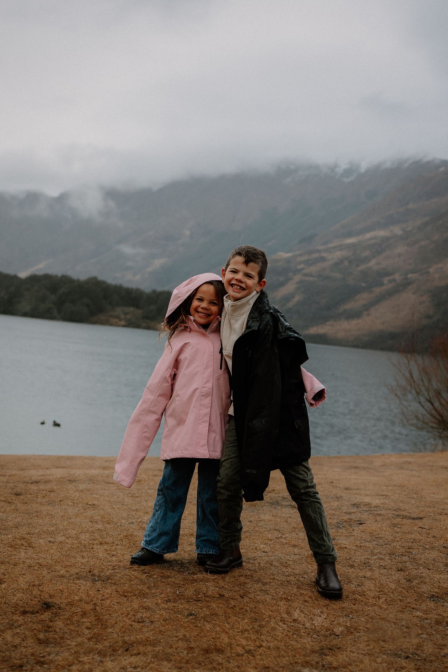 Two smiling children in rain jackets hug on a grassy lakeshore with misty mountains and an overcast sky in the background.