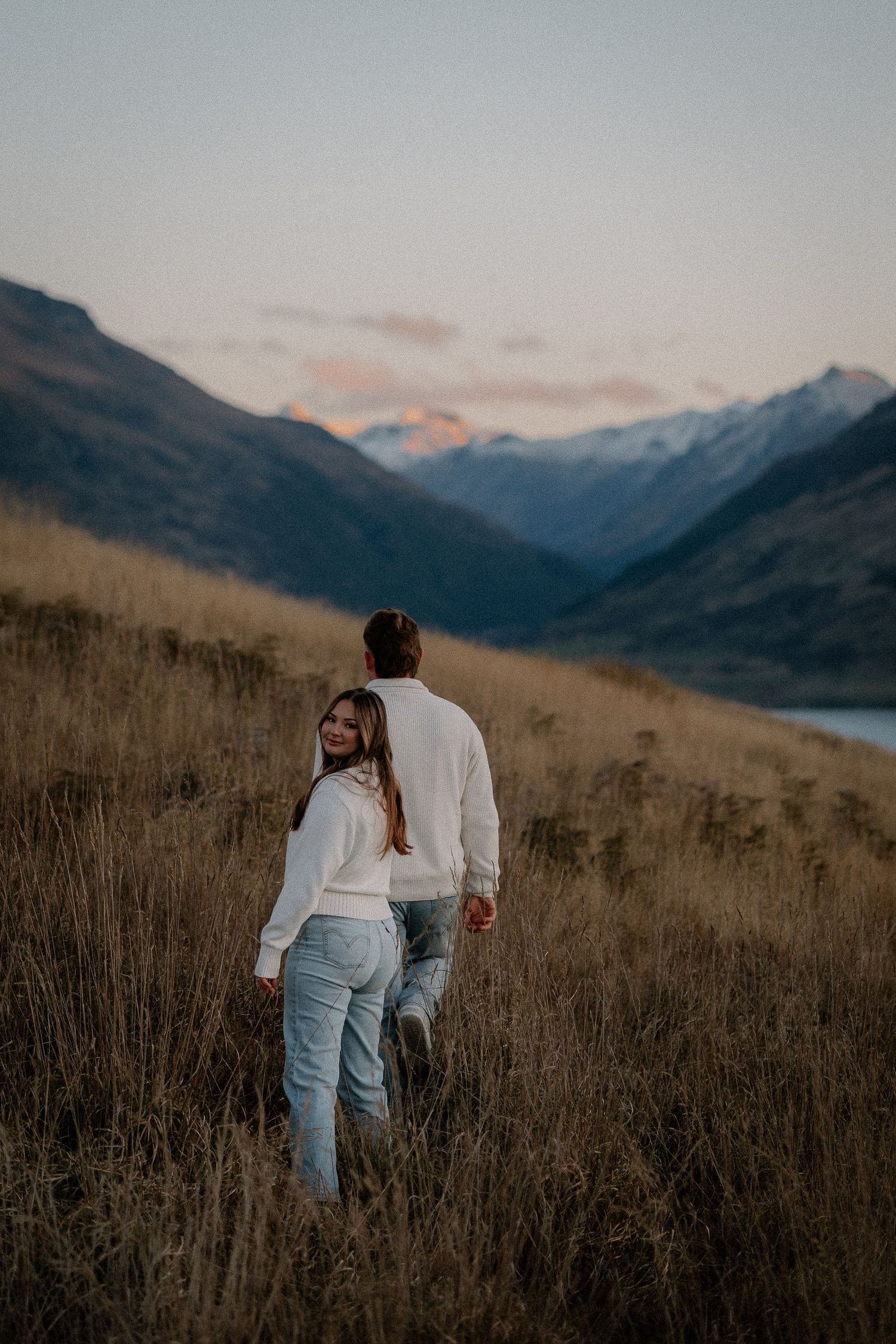Couple in white sweaters and jeans walking through golden grass on a mountain hillside at sunset, with snowy peaks and a lake behind.