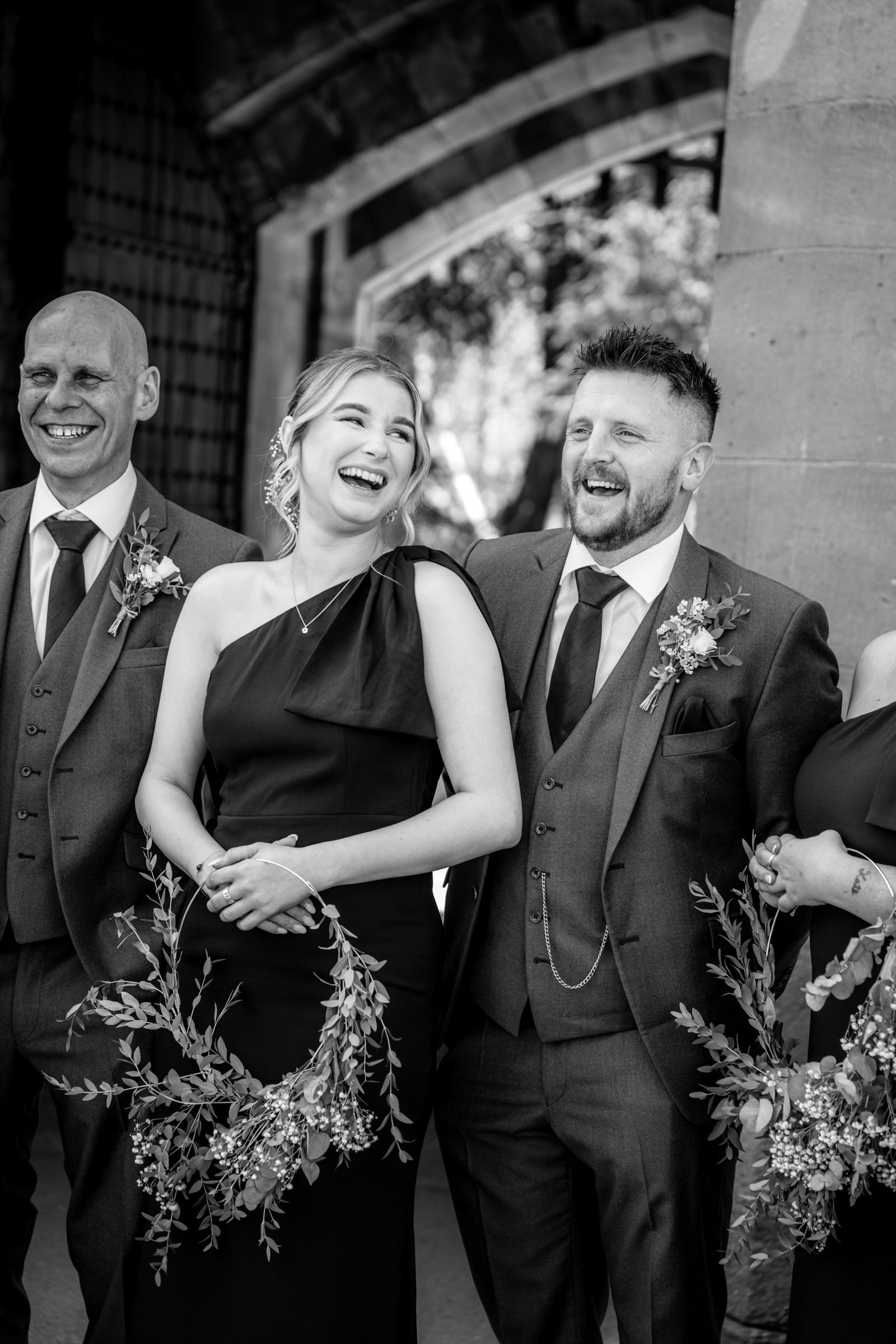 Black and white photo of laughing wedding party members in suits and a black dress, holding a floral wreath beneath a stone archway.