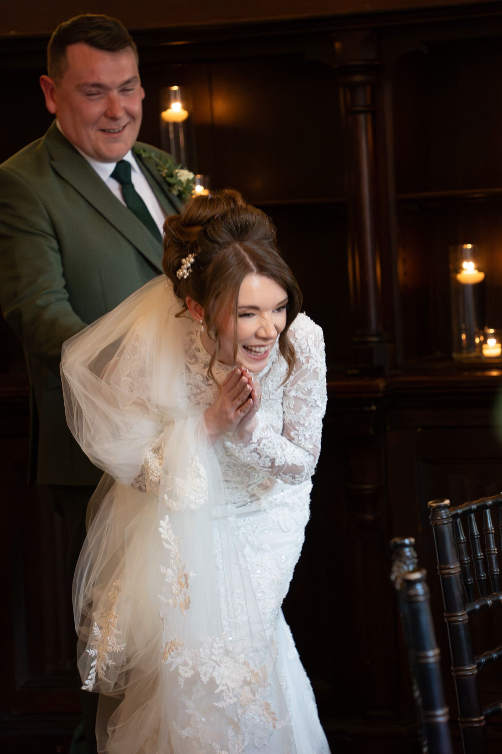 A laughing bride in a lace wedding gown and veil clasps her hands joyfully as a smiling groom in a green suit stands behind her.