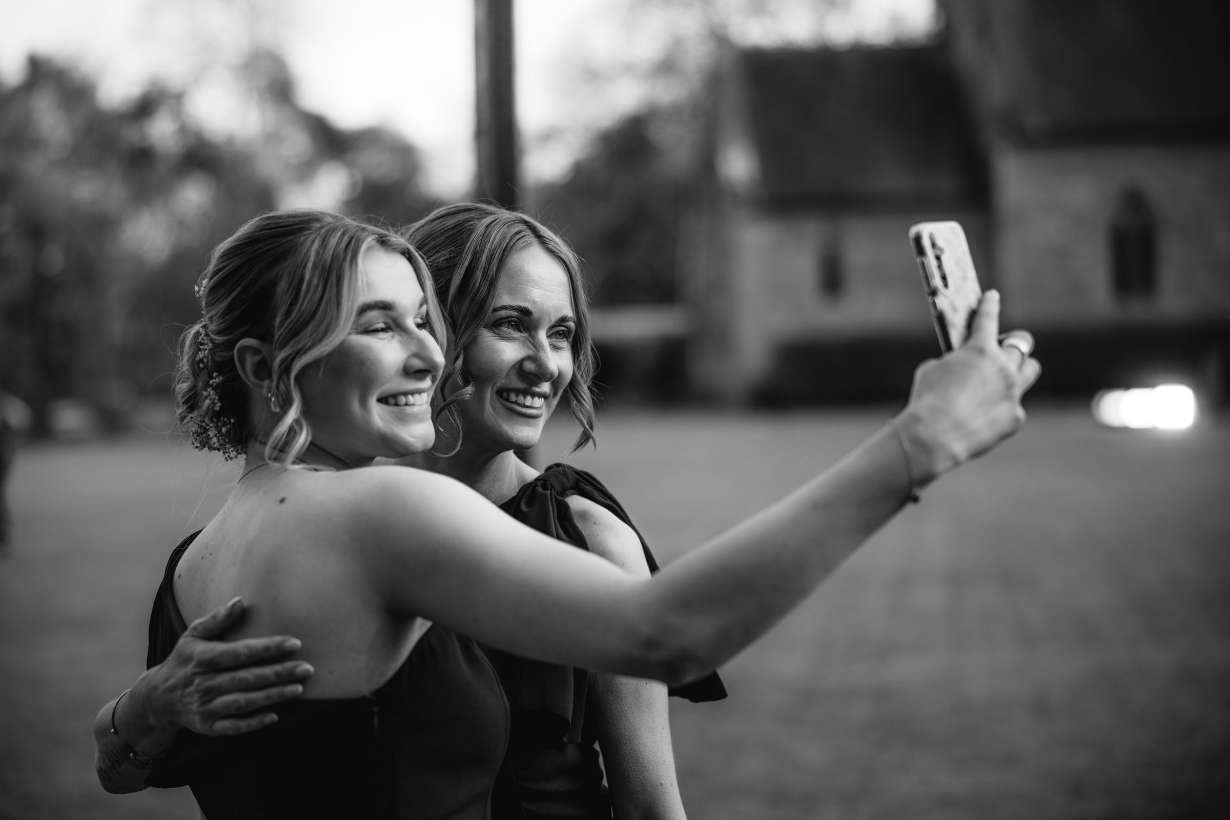 Two smiling women in dark dresses embrace and take a selfie with a smartphone on a lawn outside a stone building in black and white.
