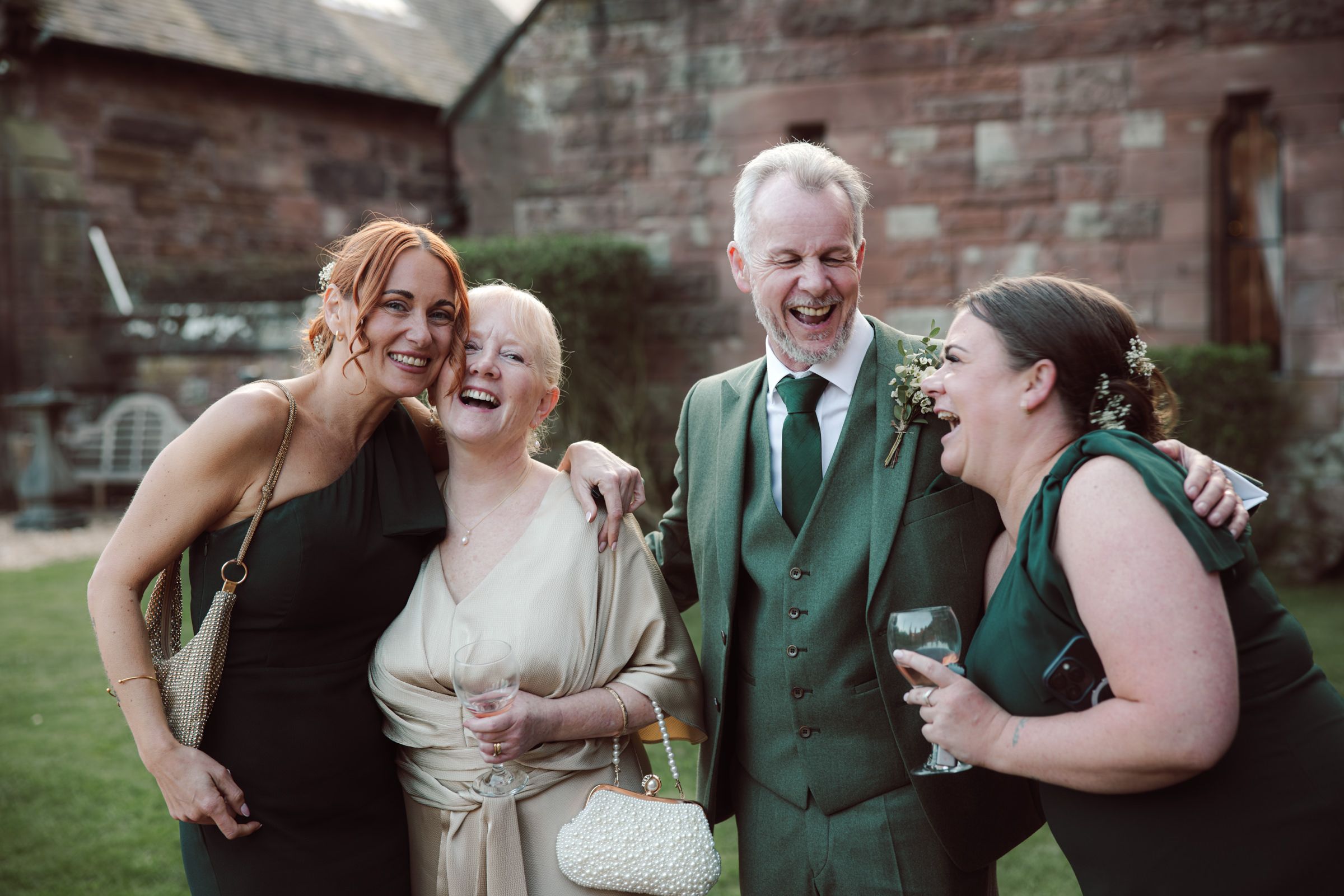Four smiling wedding guests in formal attire laugh together outdoors in front of a historic red brick building, one holding a glass.