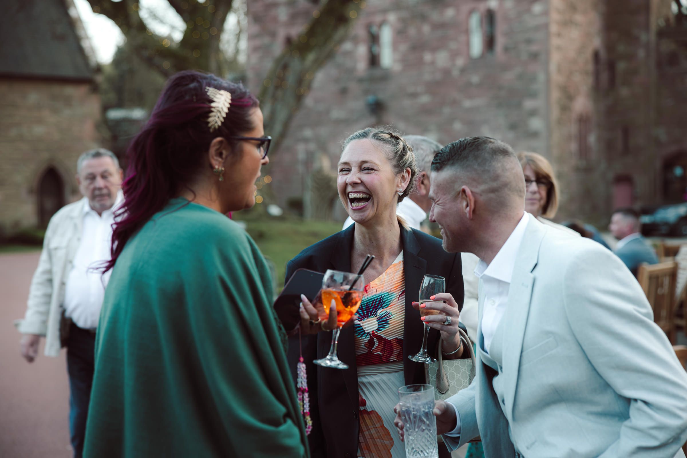 Guests laugh and chat outdoors at a stone castle venue, one woman holding a cocktail glass during a festive wedding celebration.