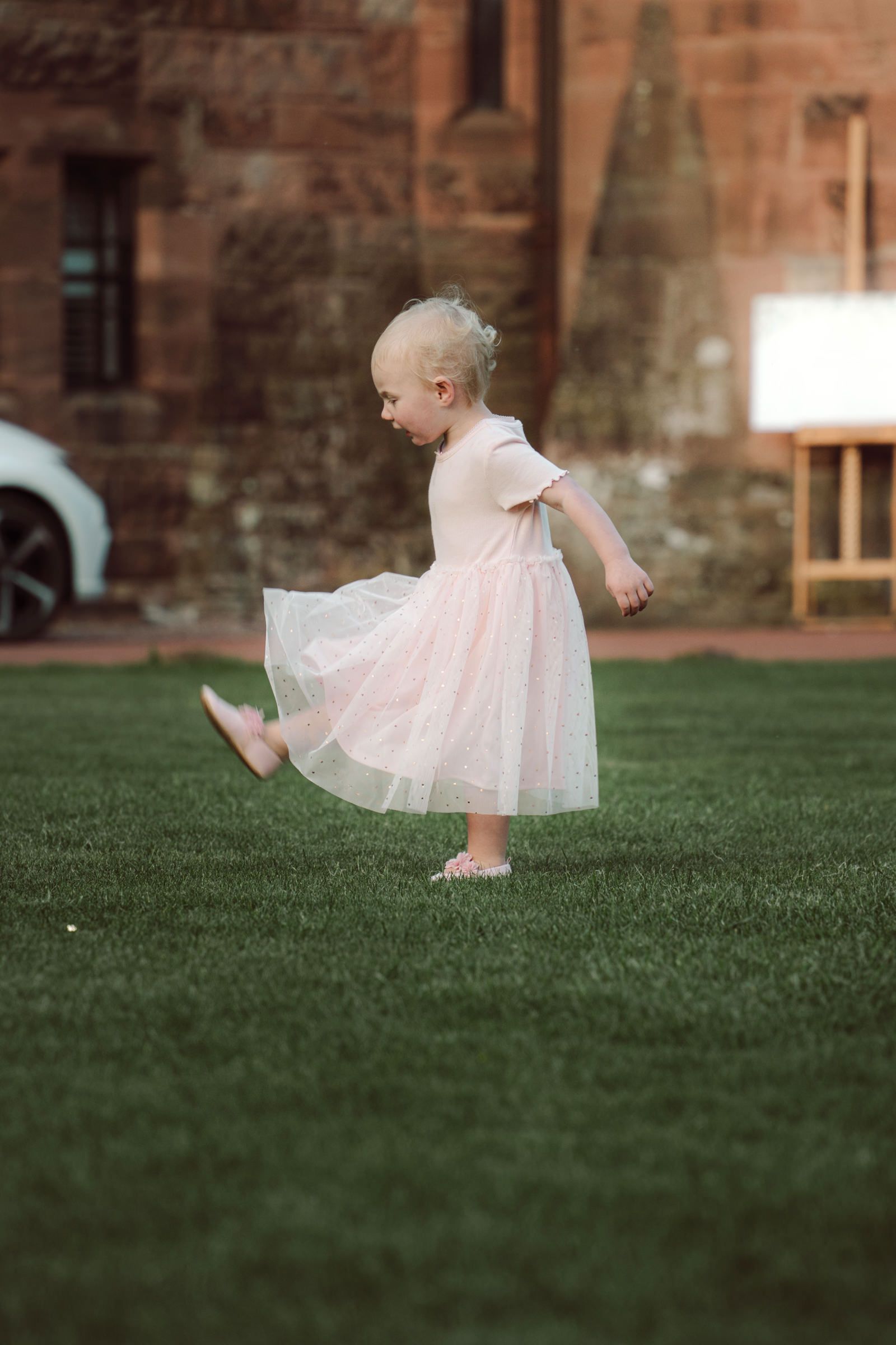 Toddler in a pink tulle dress kicks one foot up while dancing playfully on a green lawn in front of an old red sandstone building.