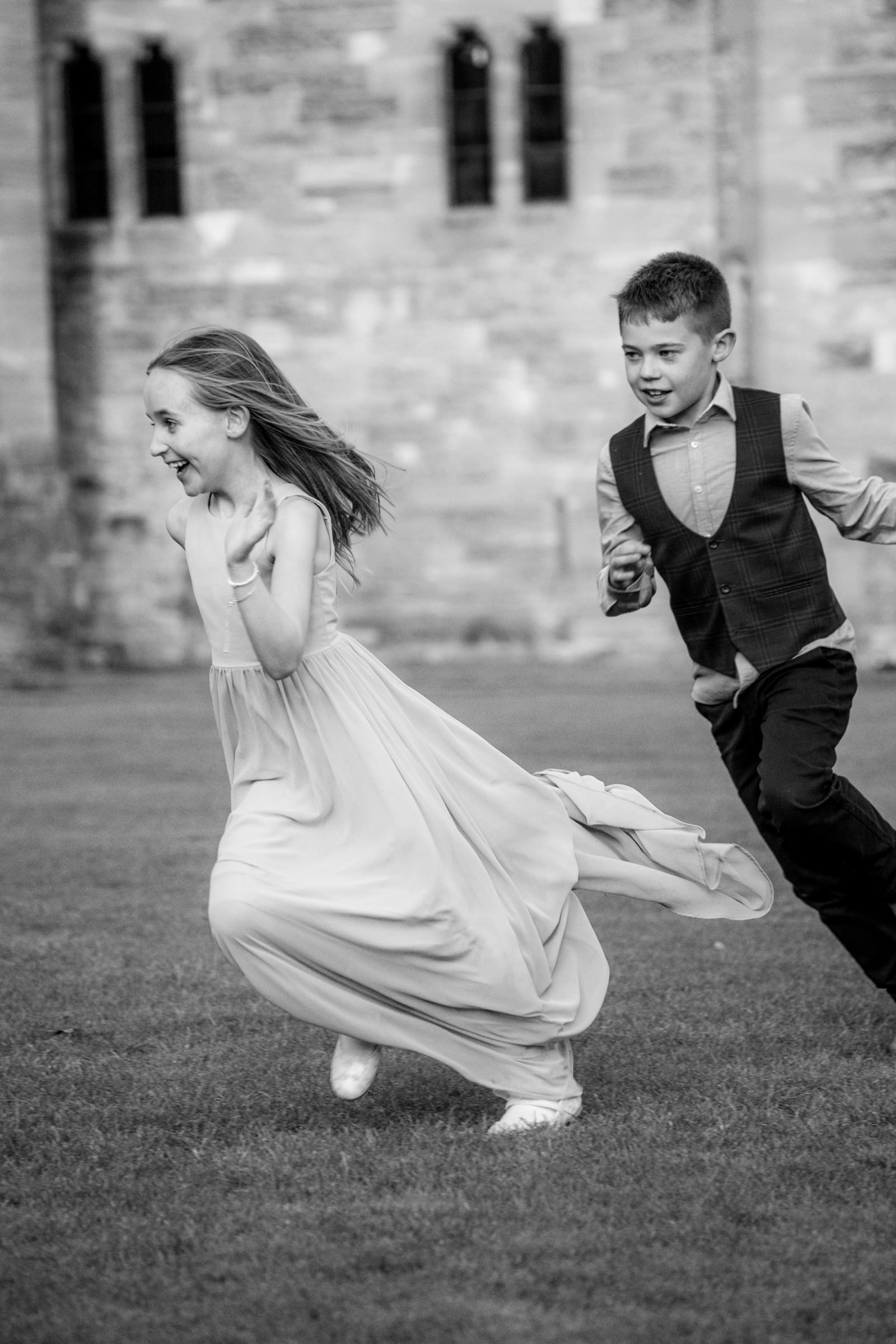 Black and white photo of a laughing girl in a flowing dress running across a lawn, chased by a boy in a vest near an old stone building.