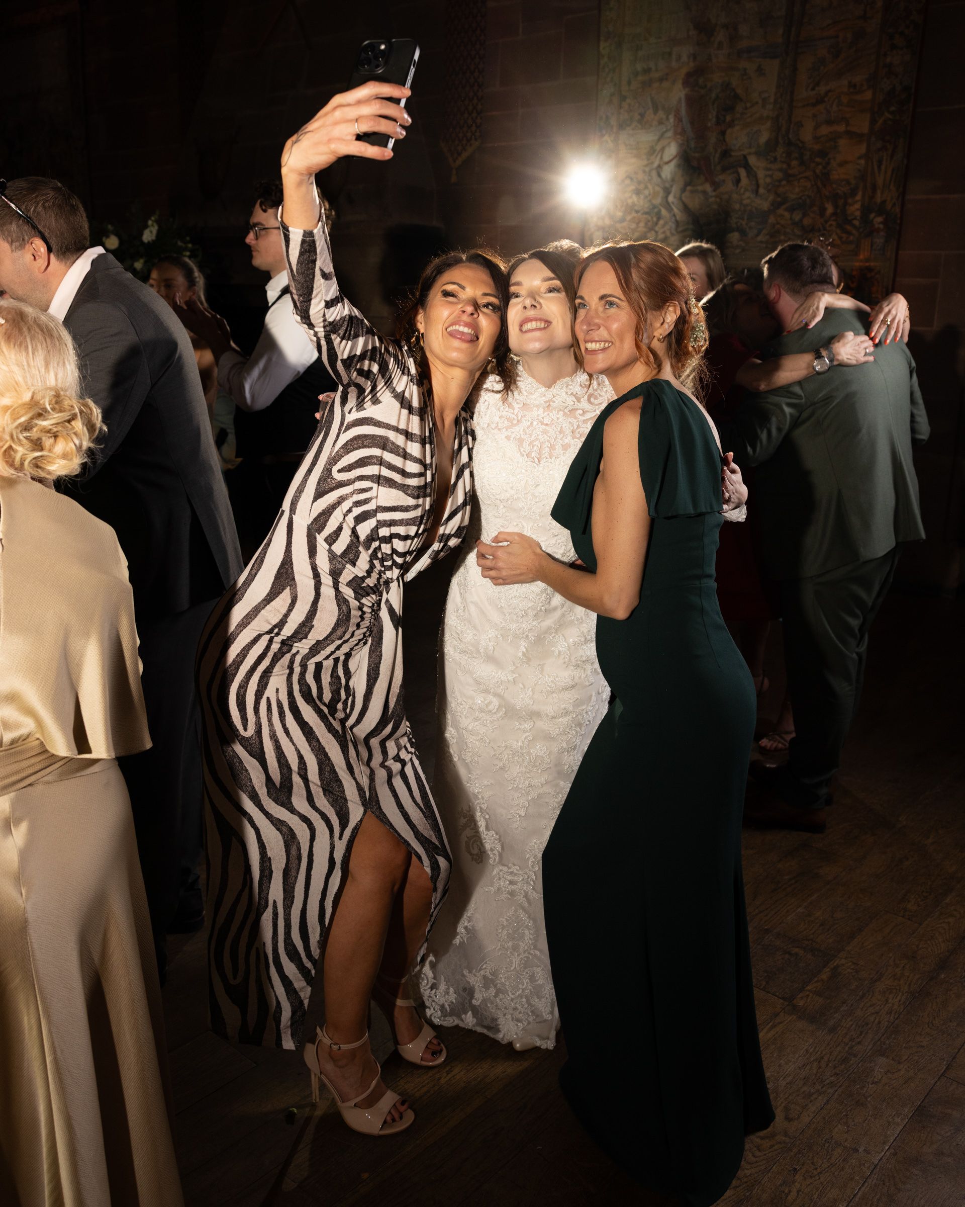 Three women in formal dresses, including a bride in white lace, pose for a joyful selfie on a crowded dance floor at a wedding reception.