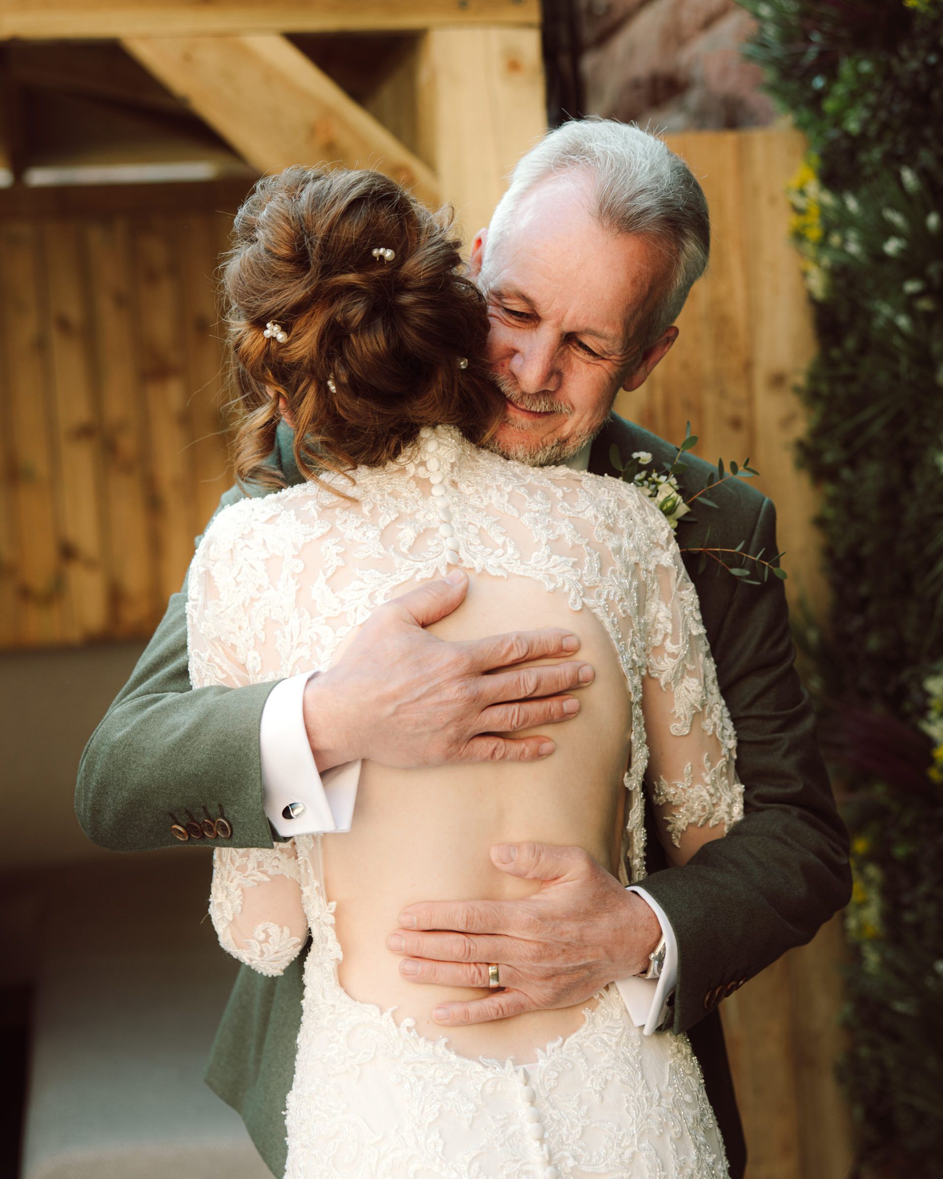 Older man in a green suit embraces a bride in a lace wedding gown with an open back and a floral updo during an emotional moment.