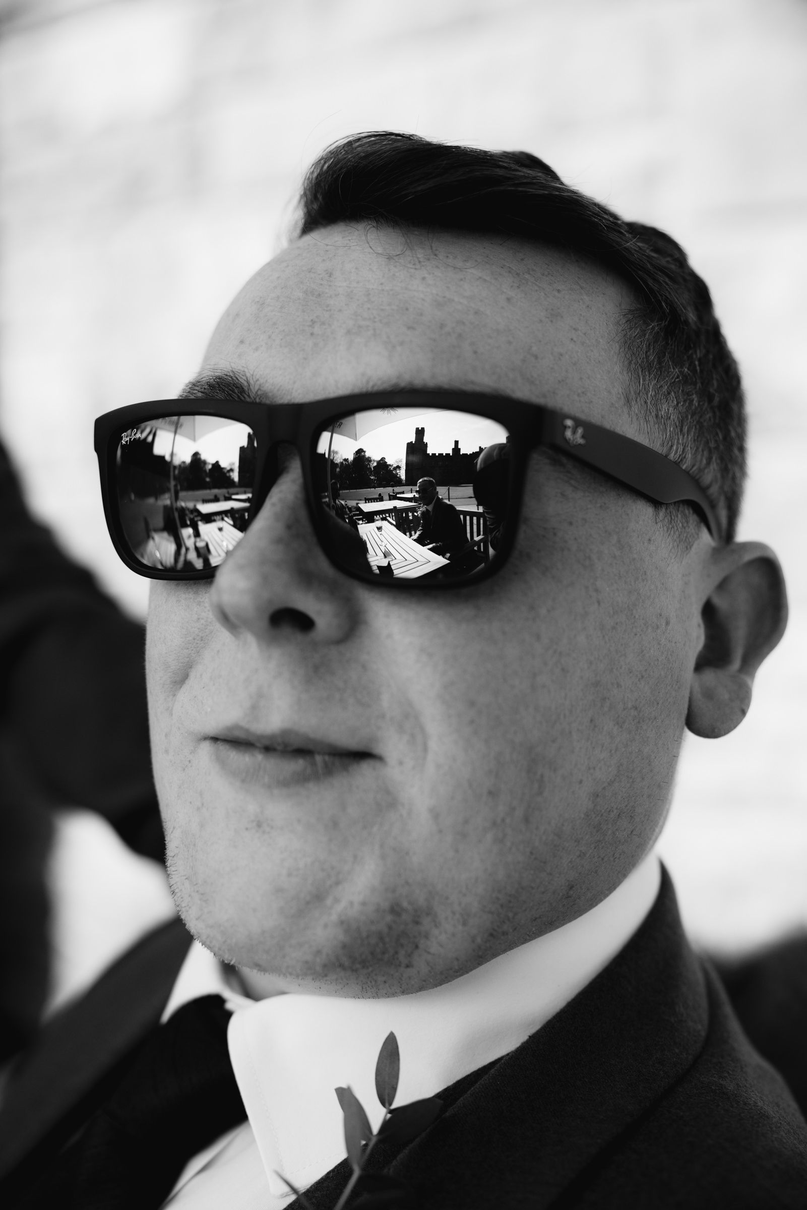 Black and white portrait of a smiling man in Ray-Ban sunglasses and a suit, with a boutonniere and buildings reflected in the lenses.