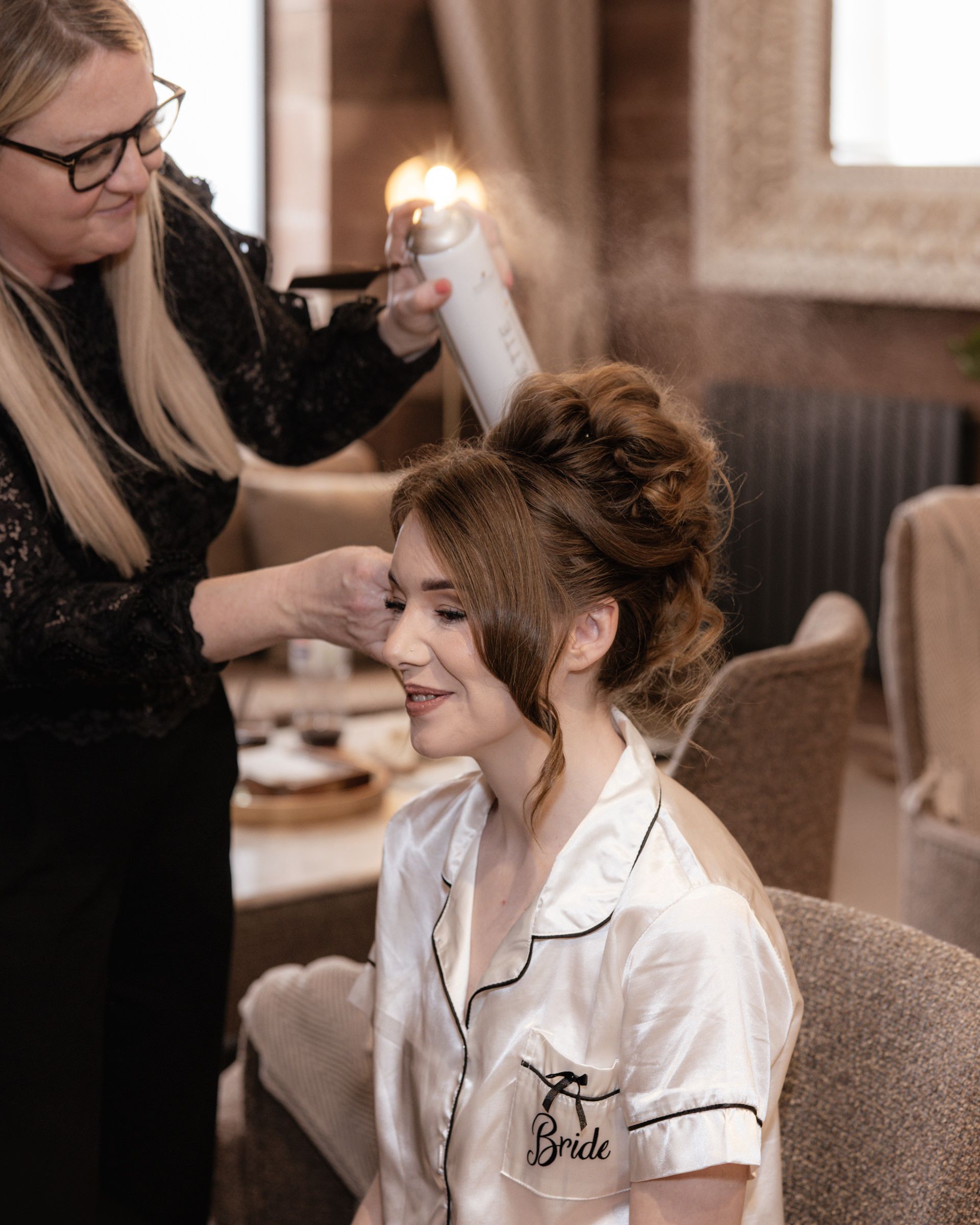 Hairstylist sprays hairspray on a bride in a white satin Bride pajama top as she gets an elegant updo before her wedding.