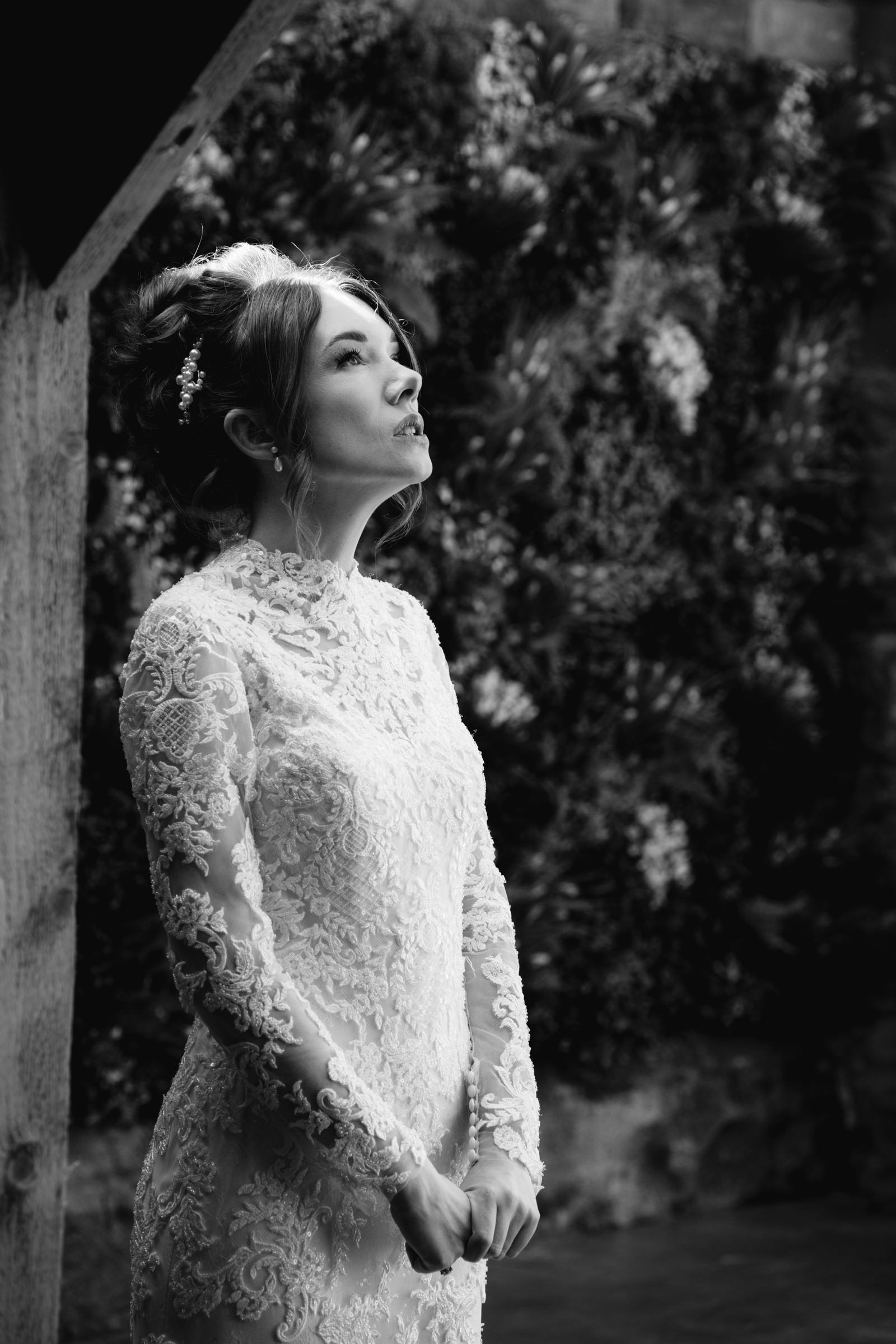 Black and white portrait of a bride in a long-sleeved lace wedding gown with an updo, gazing upward beside a wooden post and floral wall.