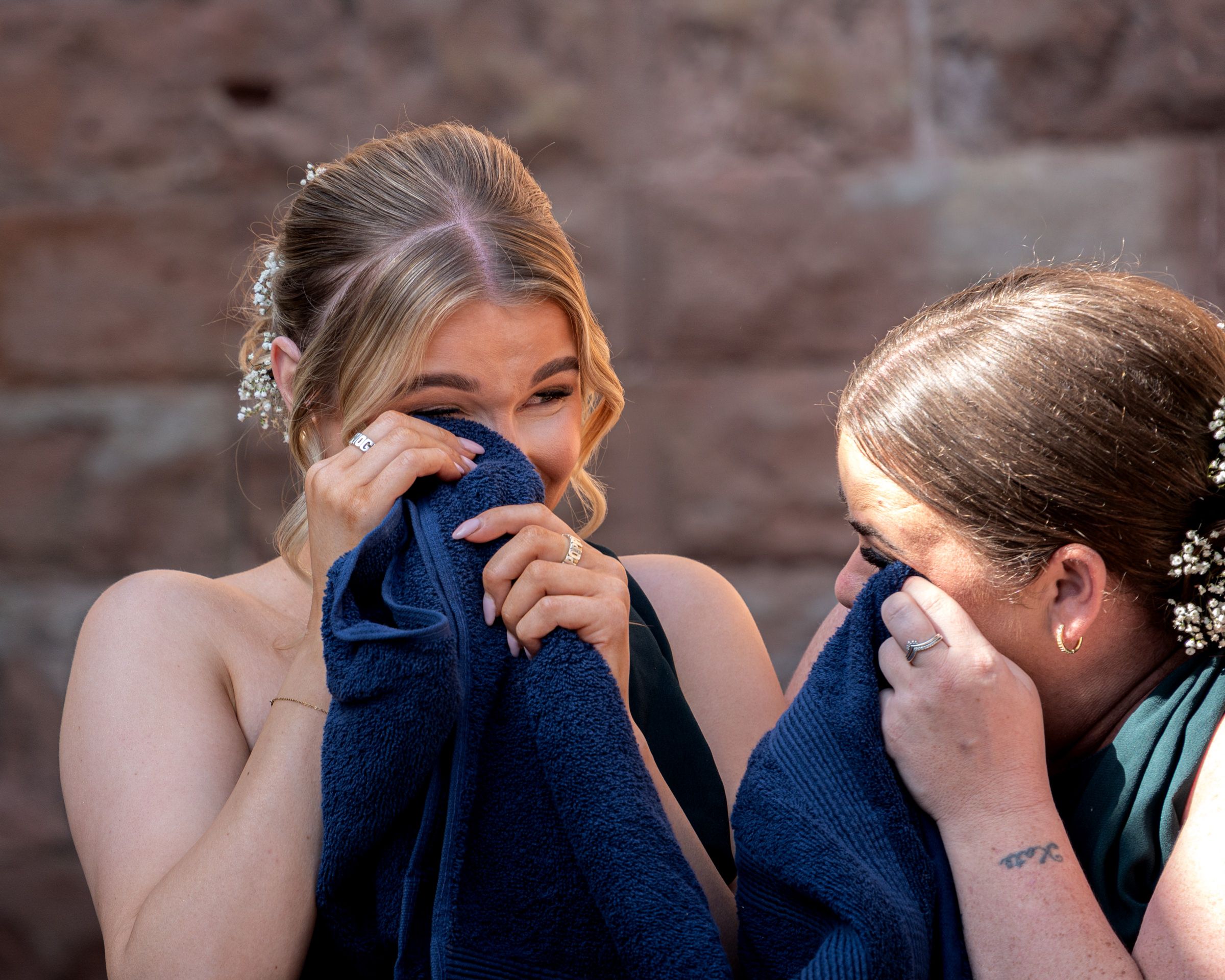 Two emotional bridesmaids in formal attire wipe tears with navy cloths while standing against a rustic stone wall.