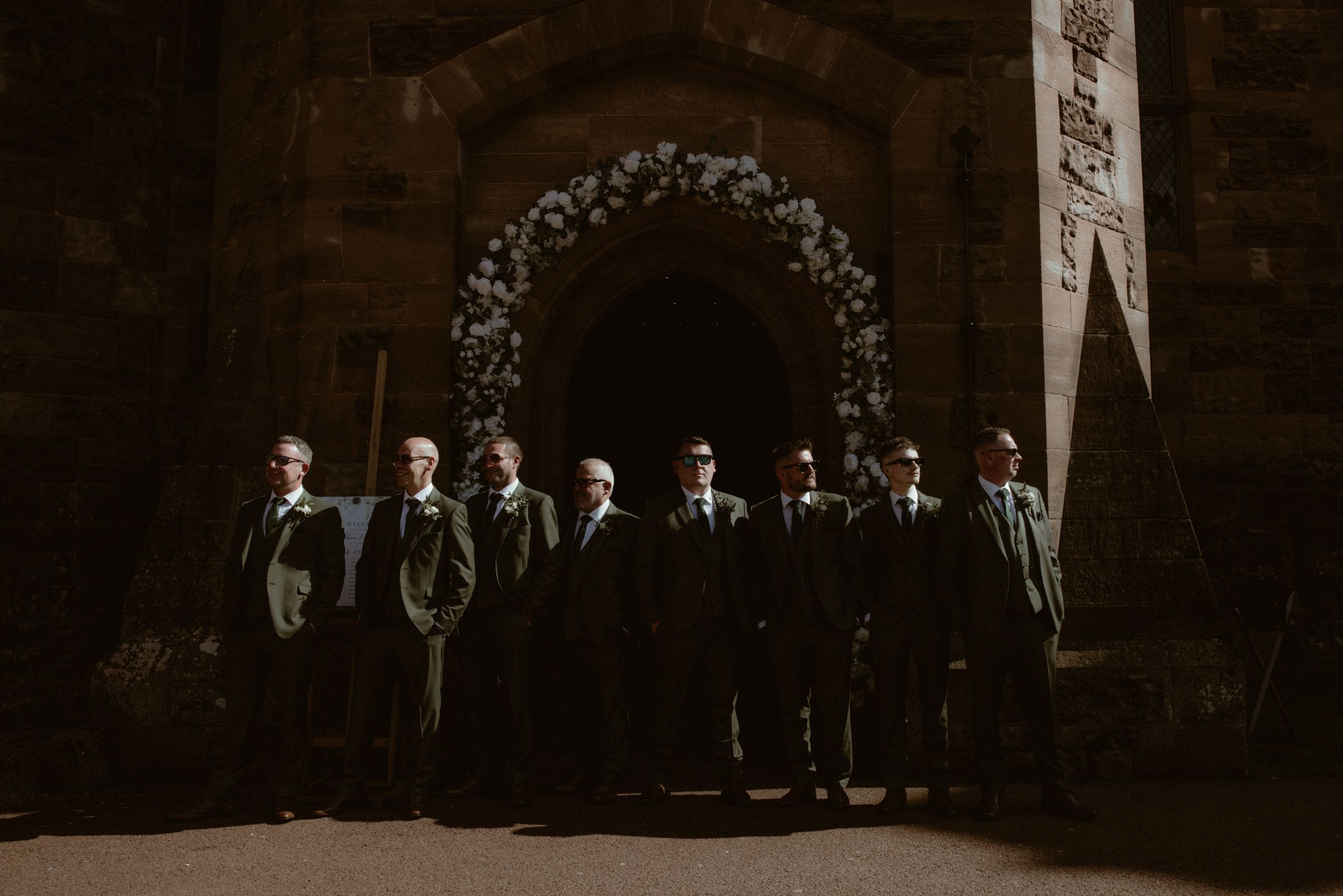 Groomsmen in dark suits stand in a row before a stone church archway decorated with a lush white floral wreath.