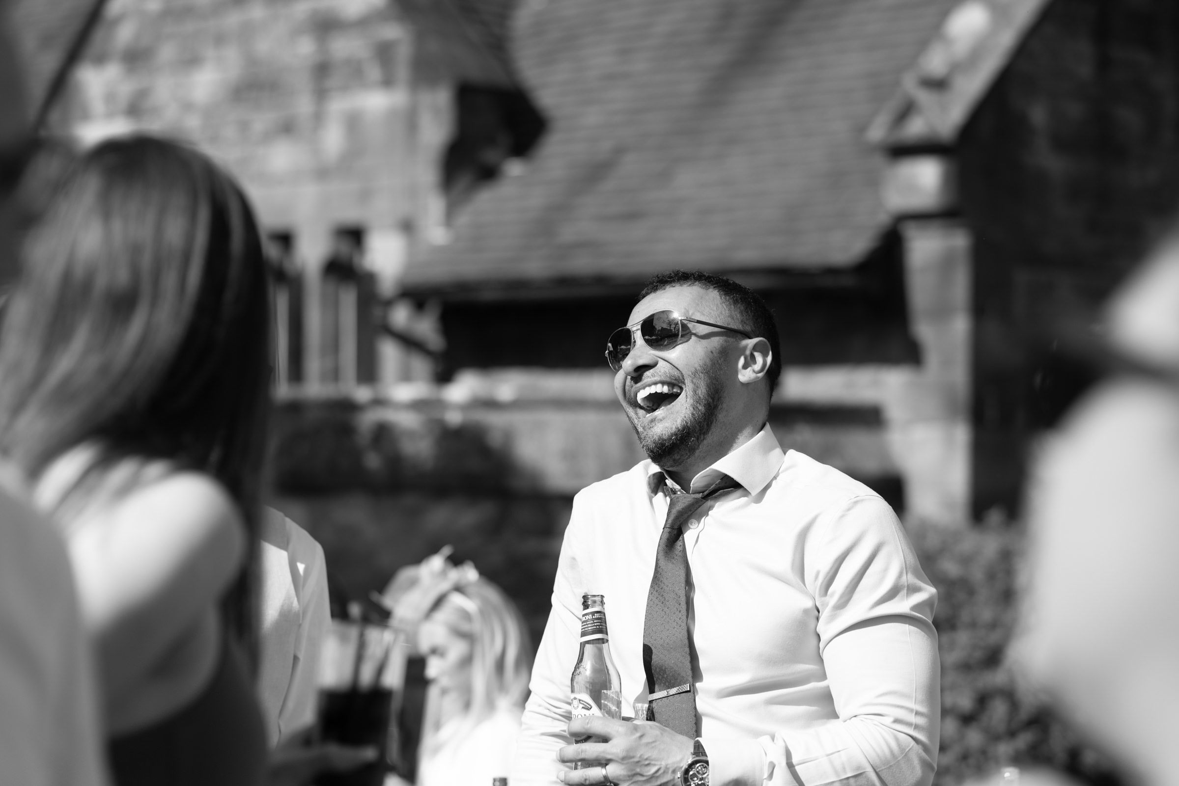 Black and white photo of a laughing man in sunglasses, shirt and loosened tie holding a bottle at an outdoor gathering.