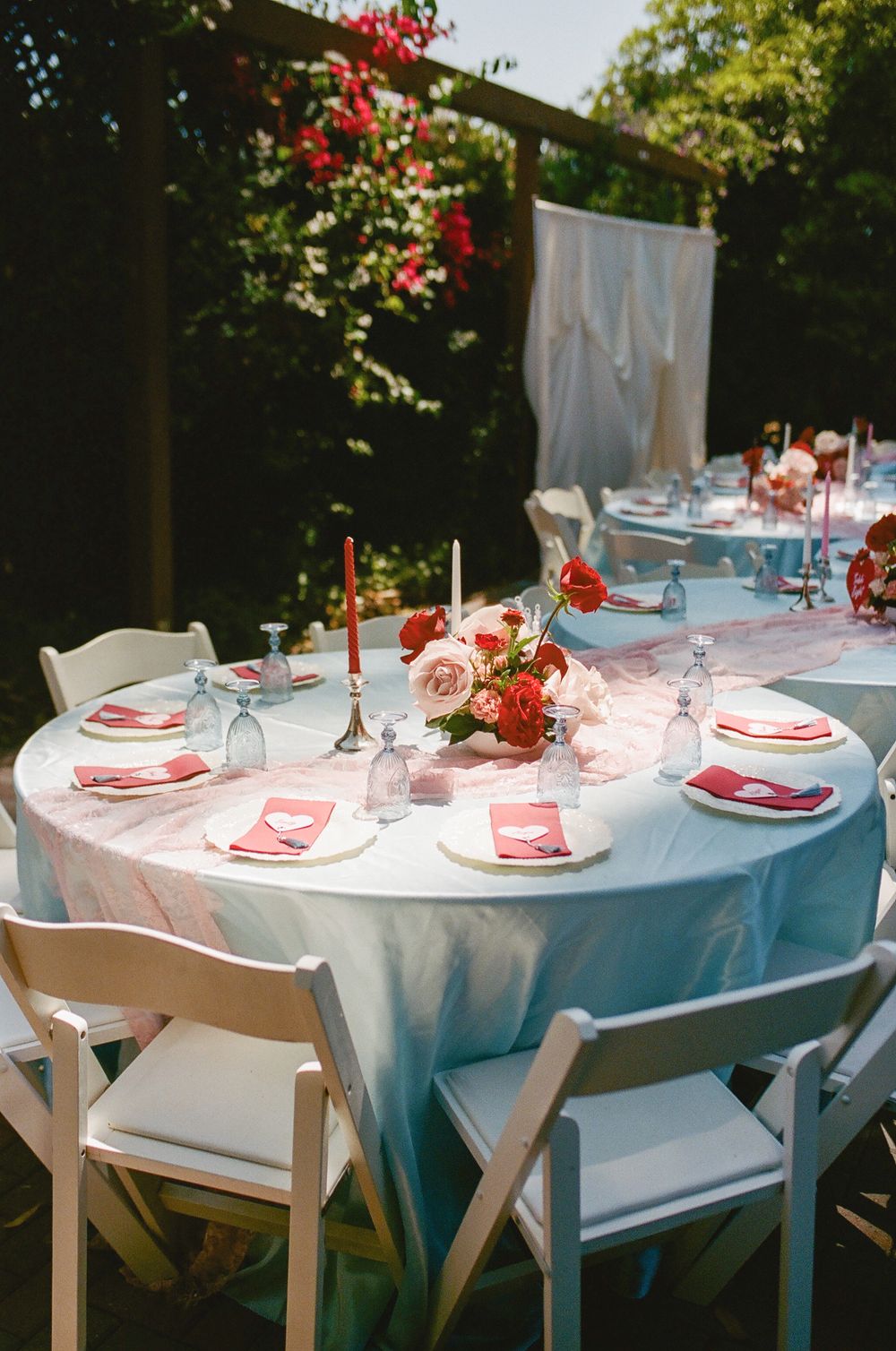 Outdoor wedding reception table with pink linens, red napkins, coral floral centerpiece, and candles beneath trees on a sunny day.