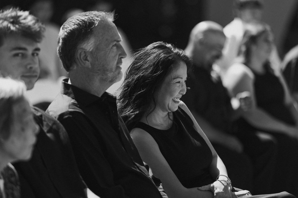 Black and white photo of a smiling woman and a man seated in an audience, watching a wedding with other attendees nearby.