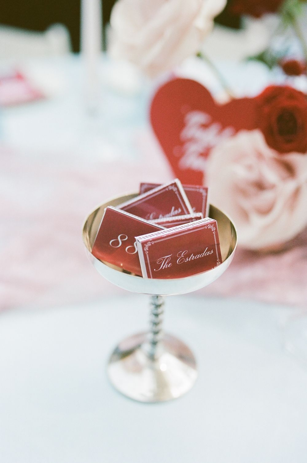Red personalized matchbooks reading The Estradas in a silver pedestal goblet beside pink roses and a red heart sign on a wedding table.