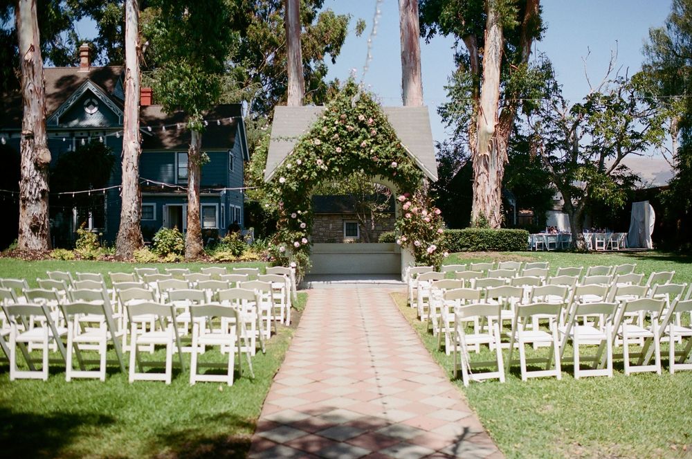 Outdoor garden wedding ceremony setup with white chairs lining a brick aisle leading to a floral arch beneath tall shady trees.