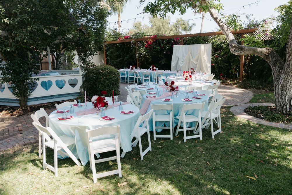 Outdoor garden wedding reception with round tables, white chairs, red floral centerpieces, and a draped ceremony arch under shady trees.