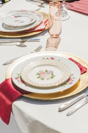 Elegant table setting with floral china plates, gold chargers, red napkins, and pink glassware on white linen.