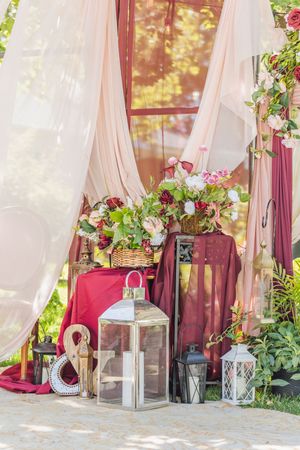 Elegant wedding decor with burgundy drapes, floral arrangements, lanterns, and an ampersand symbol outdoors.