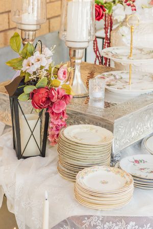 Elegant table display with stacked floral plates, candles, and pink flower arrangements on a lace-covered surface.