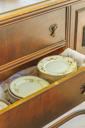 Antique wooden buffet drawer open displaying stacked vintage floral china plates wrapped in white cloth.