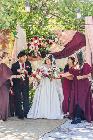 Bride and groom celebrate under floral arch with bridesmaids in burgundy dresses at outdoor wedding ceremony.
